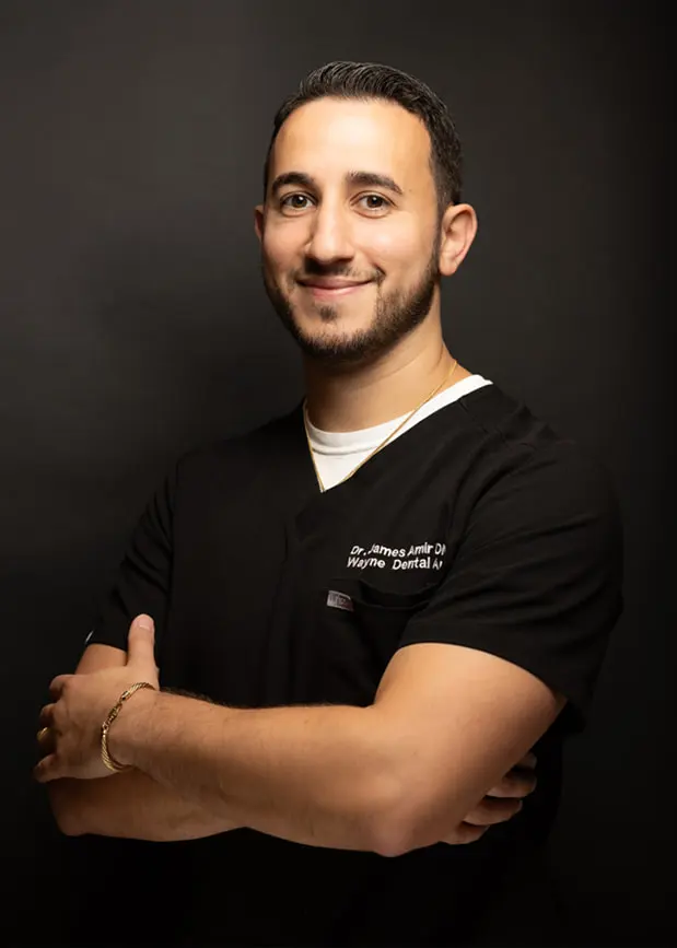 James Amir, DMD, FICOI smiling with his arms crossed wearing black scrubs with his name and the practice name embroidered on it.