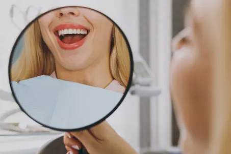 A person holding a round hand mirror that reflects their neck and dental bib in a clinical setting.