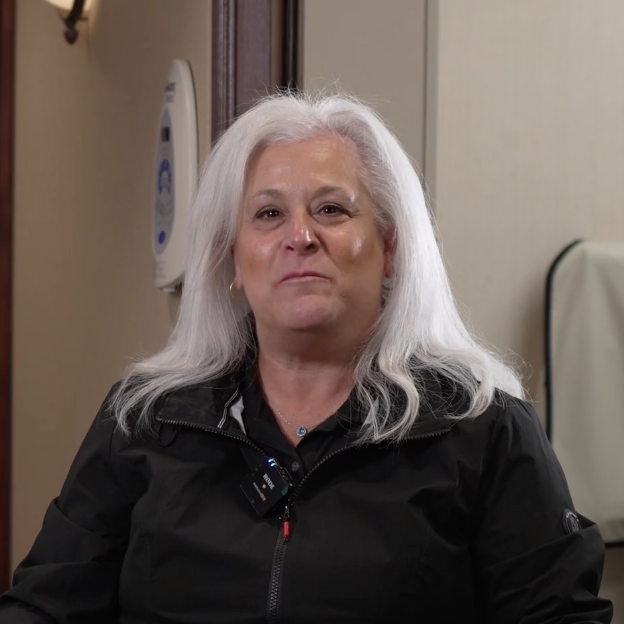 A person with long white hair, wearing a black jacket and a clipped-on microphone, sits in a dental office setting. Medical equipment and neutral-colored walls can be seen behind them.