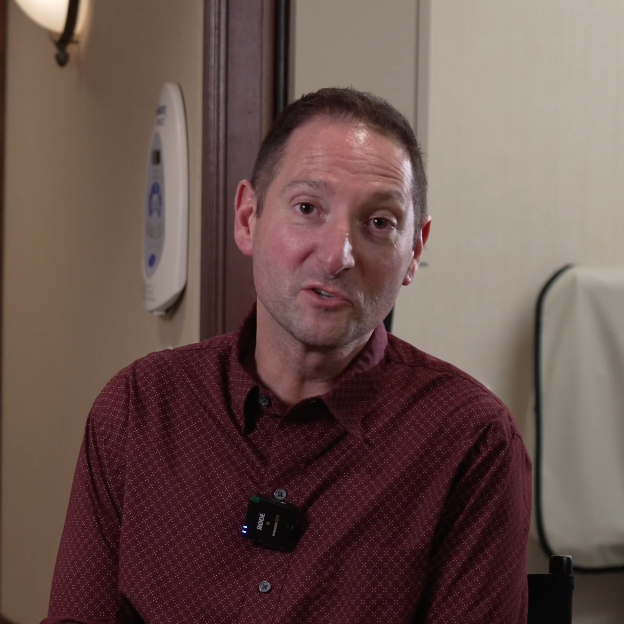 A person wearing a maroon patterned shirt sits indoors, speaking on camera with a small microphone clipped to their chest. The setting is a dentist’s office, with medical equipment visible on the wall behind them.